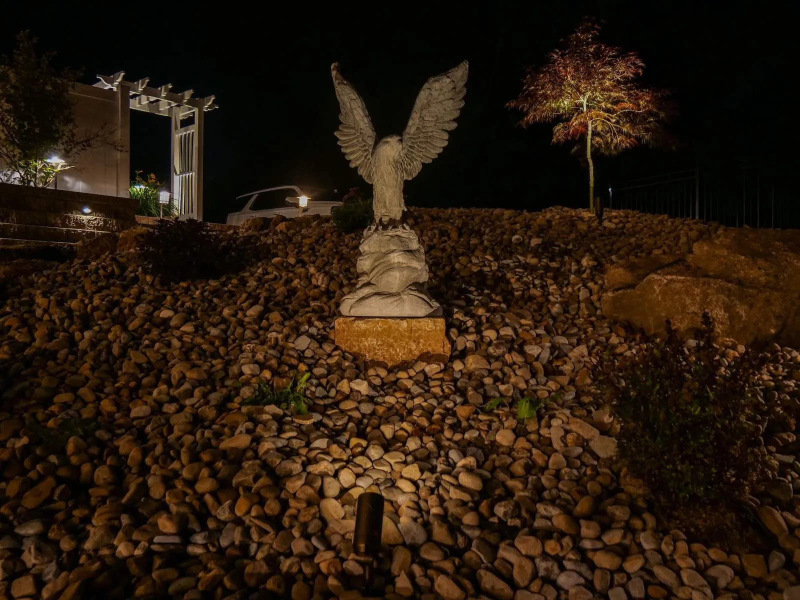 Eagle statue on a river-rock slope lit by a ground spotlight, with an uplit red-leaf tree behind it