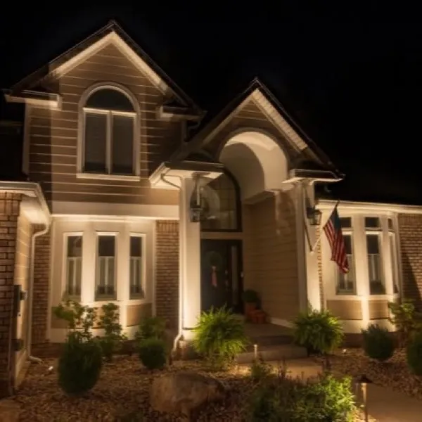 Brick and stone home lit at night with warm uplighting across the facade and garden beds